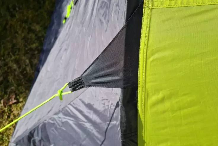 Nahaufnahme von grauem und neongrünem Stoff, einer angenähten Öse und einem neonfarbenen Spannseil; links Gras