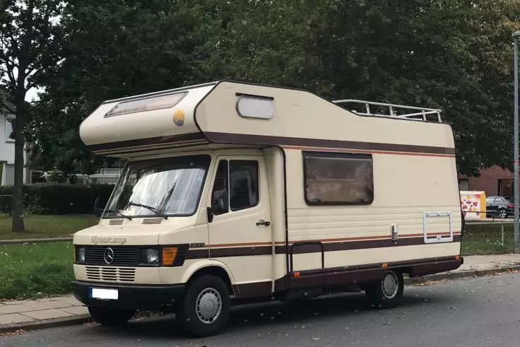 Beiges Wohnmobil mit Alkoven über der Fahrerkabine, geparkt am Straßenrand; Bäume und Rasen im Hintergrund