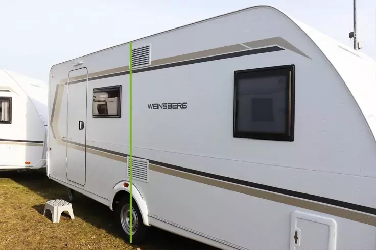 Weißer Wohnwagen mit zwei Fenstern, Eingangstür, grünem senkrechtem Streifen an der Seite, Schriftzug "WEINSBERG", kleinem Tritt davor, auf Gras
