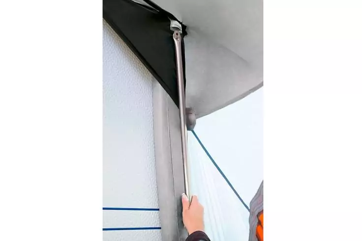 Zwei Hände halten eine vertikale Metallstange, befestigt an einer weißen Wand und einer hellen Plane; grünes Seil verläuft daneben; rechts ein orangefarbener Ärmel