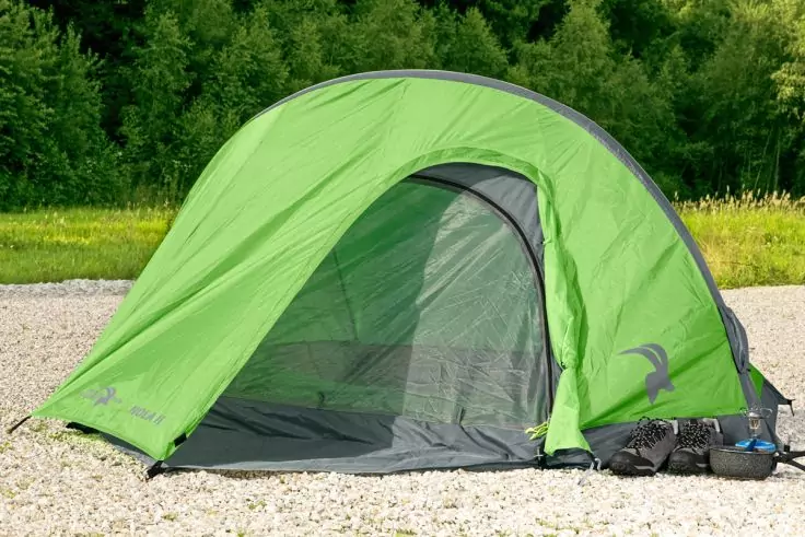 Grünes Zelt mit offenem Innenzelt auf Kies, schwarze Schuhe und Rucksack rechts, Grasstreifen und Bäume im Hintergrund