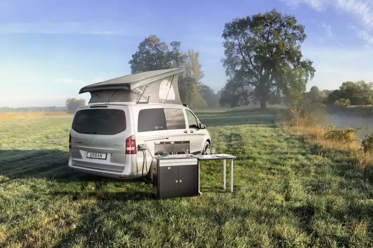 Silberner Camper mit aufgeklapptem Aufstelldach auf einer Wiese, daneben ein Klapptisch und ein schwarzes Gerät, im Hintergrund Bäume und blauer Himmel