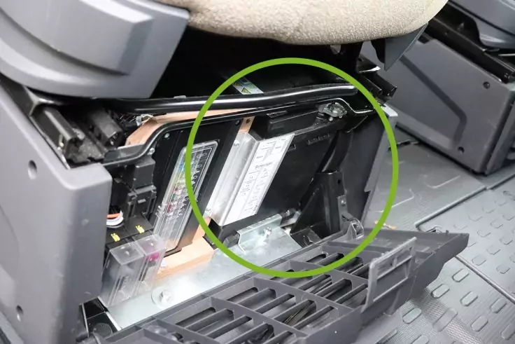 Geöffnete Verkleidung unter einem Fahrzeugsitz; freigelegtes Metallgehäuse mit Etikett, sichtbare Kabel, abgeklappte Kunststoffabdeckung auf geriffeltem Boden; grüner Kreis um den Bereich