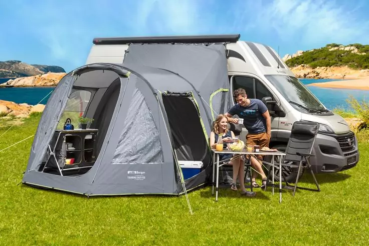 Grauer Camper mit angebautem Vorzelt auf grünem Gras, zwei Personen sitzen an einem Campingtisch vor dem Vorzelt; im Hintergrund Felsen, Meer und blauer Himmel