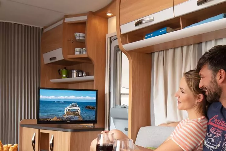 Innenraum eines Wohnmobils mit Holzschränken und Regalen, Fernseher zeigt ein Boot auf dem Meer, Regal mit Tassen, eine Frau und ein Mann sitzen nebeneinander und halten Weingläser