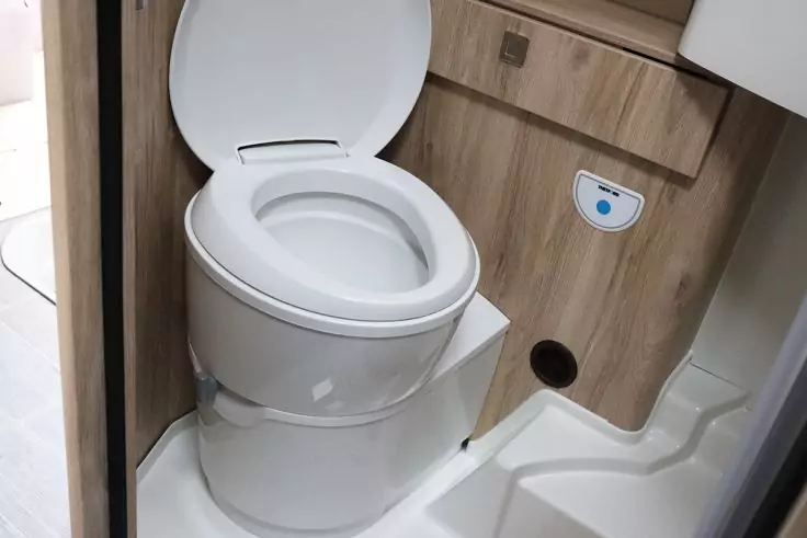 Weißes Toilettenbecken mit geöffnetem Sitz, daneben Holzverkleidung mit rundem Loch und kleinem weißem Panel mit blauem Punkt