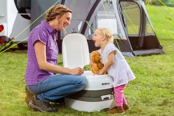 Erwachsene kniet auf Gras vor einem kleinen Kind, das auf einer tragbaren Campingtoilette steht und einen Teddybären hält; Wohnmobil und Zelt im Hintergrund