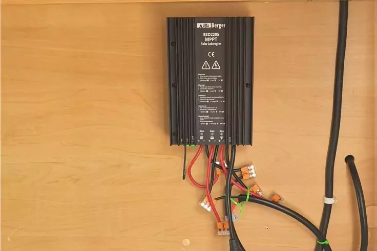 Schwarzes elektronisches Gerät mit Aufschriften, an einer Holzplatte befestigt, mehrere farbige Kabel und Steckverbinder darunter