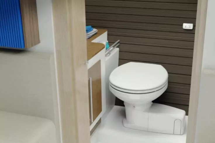 Weißes geschlossenes WC vor brauner Wand mit horizontalen Lamellen; weißes Waschbeckenmöbel mit offenem Fach, kleine Flasche und gefaltetes Handtuch auf der Ablage; heller Boden; Türrahmen sichtbar