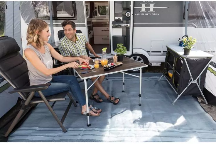 Zwei Personen sitzen an einem Campingtisch vor einem Wohnwagen; Teller, Gläser und Campingmöbel sichtbar