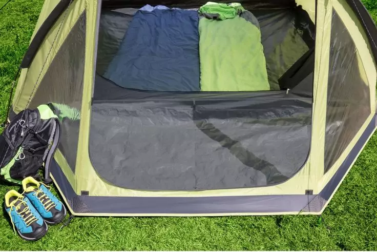 Innenraum eines grünen Zelts mit zwei Schlafsäcken (blau und hellgrün) auf grauem Boden, am Eingang ein Rucksack und ein Paar blaue Sportschuhe auf Gras