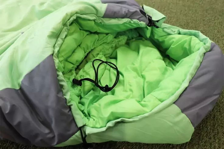 Grün-grauer geöffnet liegender Schlafsack mit schwarzer Kordel im Inneren auf grünem Untergrund
