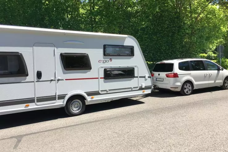 Weißer Pkw mit angehängtem weißen Wohnwagen, geparkt am Straßenrand vor grünen Bäumen