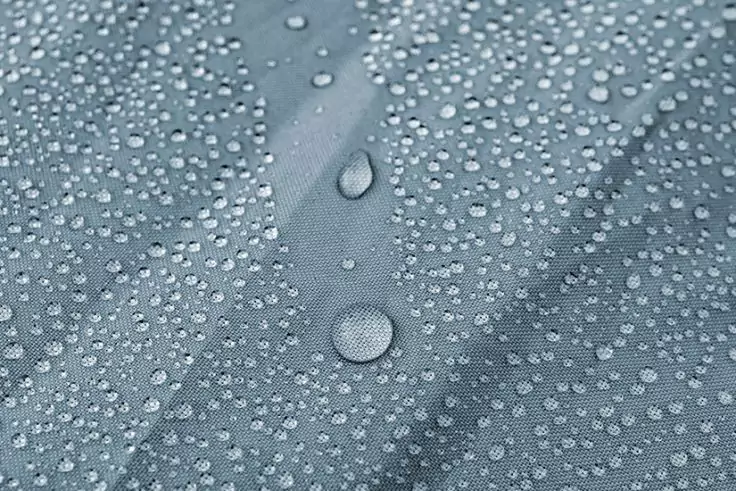 Nahaufnahme eines graublauen Stoffs mit vielen Wassertröpfchen, mehrere größere Tröpfchen entlang einer diagonalen Naht