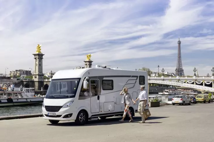 Weißes Wohnmobil an einer Uferpromenade, zwei Personen daneben, Brücke mit goldenen Figuren und Eiffelturm im Hintergrund