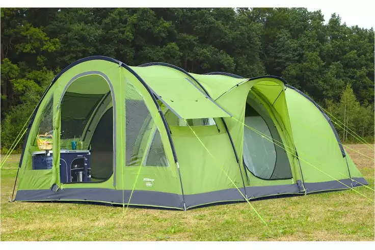 Grünes Tunnelzelt mit drei Bögen auf einer Wiese, vorderer Eingang offen, Innenraum sichtbar mit blauem Stuhl und Tisch, abgespannte Abspannleinen, Bäume im Hintergrund
