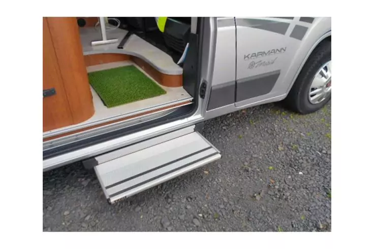 Offene Seitentür eines Fahrzeugs mit ausgeklapptem Metalltritt, grünem Fußmatte im Innenraum, Holzschwelle und grauem Schotterboden