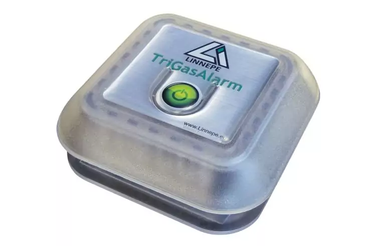 Quadratisches, transluzentes Kunststoffgerät mit abgerundeten Ecken. Auf der Oberseite ein Etikett mit dreieckigem Logo, dem Schriftzug 'TriggaAlarm' und einer grünen, runden Taste