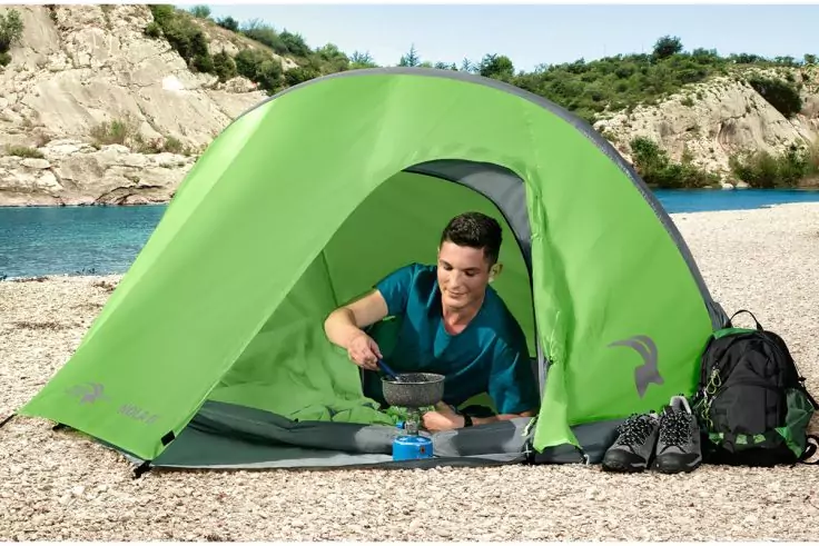 Person in grünem Zelt auf Kiesstrand, über einer Pfanne hockend; Rucksack und Wanderschuhe neben dem Zelt; Meer und Felsen im Hintergrund