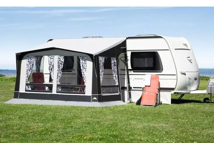 Weißer Wohnwagen mit angebautem Vorzelt auf grünem Rasen, gemusterte Vorhänge in den Fenstern, roter Klappliegestuhl am Eingang, Meer im Hintergrund, klarer Himmel