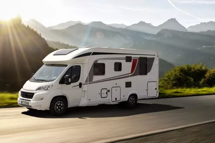 Weißes Wohnmobil auf einer Straße vor Bergen und Sonnenstrahlen