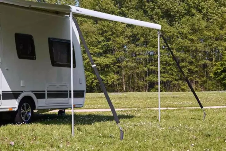 Weißer Wohnwagen mit ausgefahrener Markise, zwei Stützgestänge mit Abspannseilen auf einer Wiese, Baumreihe im Hintergrund