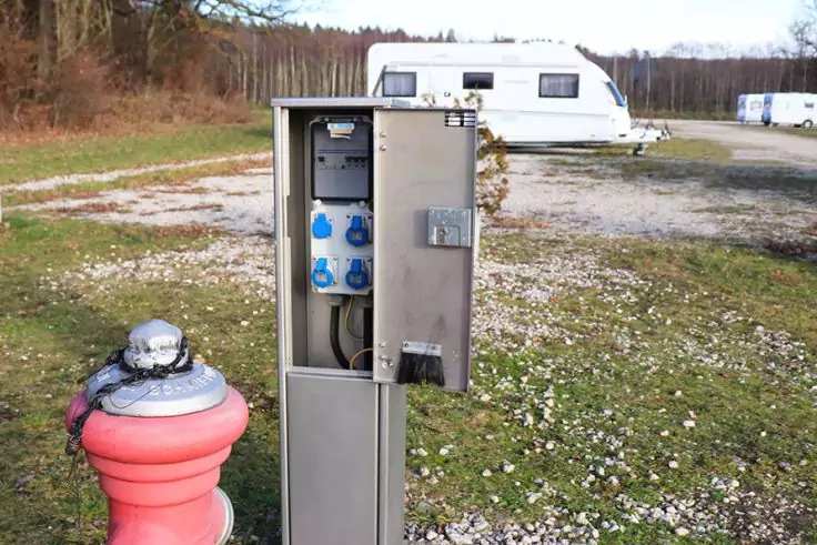 Offene Metall-Säule mit blauen Steckdosen und sichtbaren Kabeln, im Vordergrund roter Hydrant mit silberner Abdeckung, im Hintergrund weiße Wohnwagen und Baumreihe