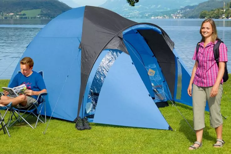 Blaues Zelt auf Gras am Seeufer, ein Mann sitzt auf einem Campingstuhl und liest eine Zeitschrift, eine Frau steht mit Rucksack; See und Hügel im Hintergrund