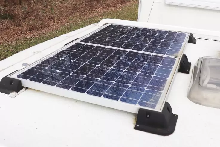 Rechteckiges Solarmodul mit blauen Zellen auf weißem Dach, befestigt mit schwarzen Halterungen; rechts eine Dachluke, im Hintergrund Gras