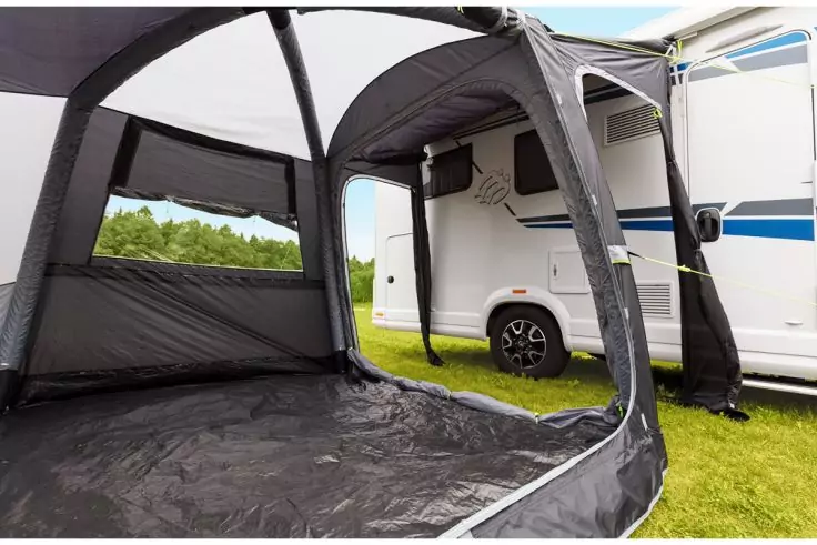 Aufblasbares Vorzelt an weißem Wohnmobil, offener Eingang, schwarzer Boden, Rasen und Bäume im Hintergrund