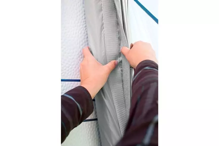 Zwei Hände halten einen grauen Stoff mit sichtbarem Reißverschluss; heller Hintergrund mit blauen Streifen