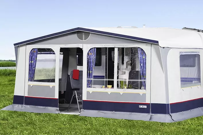 Weiß-blaues Vorzelt neben einem Wohnwagen auf Gras; große Fenster mit Vorhängen, offener Eingang mit kleinem Tritthocker, Felder und Himmel im Hintergrund