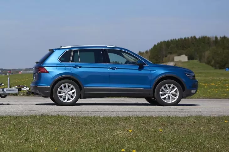 Blauer SUV in Seitenansicht auf einer Straße mit kleinem Anhänger, Gras im Vordergrund, Bäume und Gebäude im Hintergrund, blauer Himmel