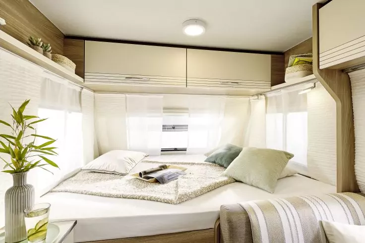 Schlafbereich eines Wohnmobils mit Doppelbett, zwei Kissen, Tagesdecke, offenes Buch auf dem Bett, Hängeschränken, Fenster mit weißen Vorhängen, Pflanze in Vase auf einer Ablage