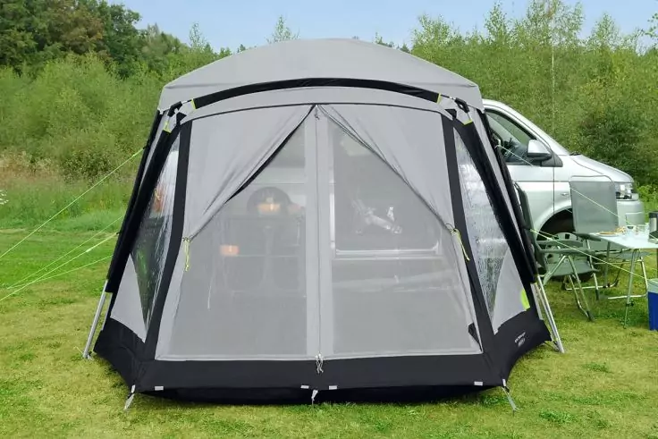 Graues Vorzelt mit feinmaschigem Netz vorne auf Rasen; rechts ein weißer Campingbus, daneben Tisch und Klappstühle; im Hintergrund Bäume