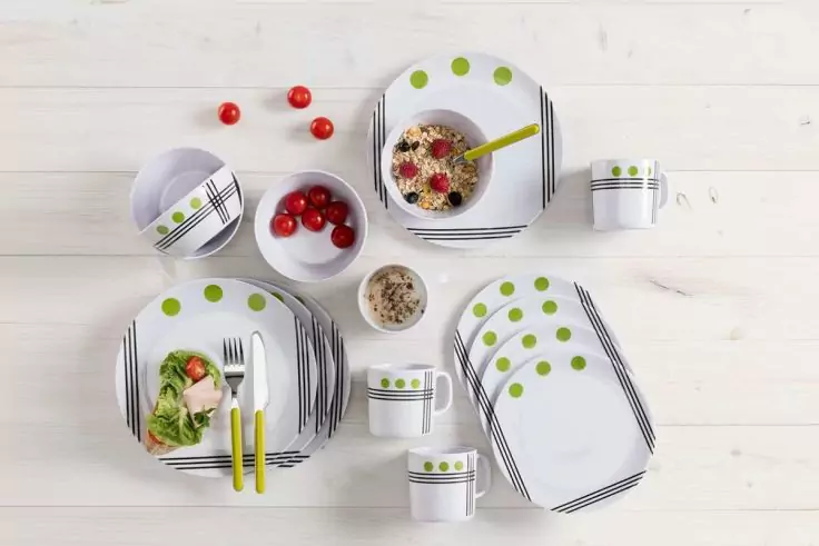 Weiße Geschirrserie mit grünen Punkten und schwarzen Streifen auf hellem Holzhintergrund; Teller, Schalen, Tassen, Besteck mit gelben Griffen, Schale mit Flocken, roten Beeren und Löffel, Teller mit Salat, verstreute rote Kirschen