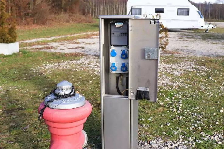 Offener grauer Metallkasten mit blauen Steckdosen; davor ein rotes Standrohr mit rundem silbernem Deckel; im Hintergrund ein weißer Wohnwagen auf Kies, Gras und Bäume