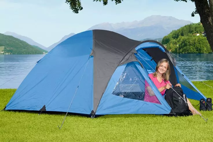 Blaues Zelt auf Gras am See; Frau sitzt im Eingangsbereich und hält einen Rucksack; Berge und Bäume im Hintergrund; Schuhe neben dem Zelt