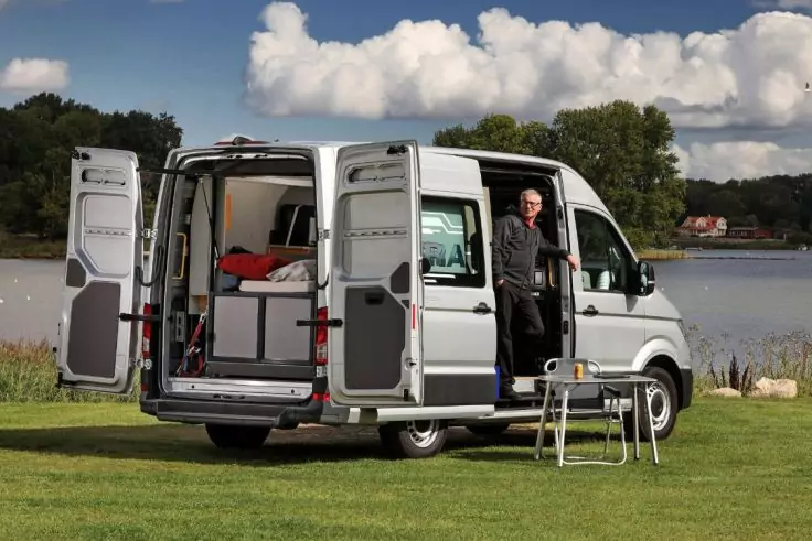 Silberner Camper-Van auf Wiese am See, Heck- und Seitentür geöffnet, Innenraum mit Bett und Schränken sichtbar, ein Mann steht in der Seitentür, kleiner Klapptisch und zwei Stühle davor, See, Bäume und Wolken im Hintergrund