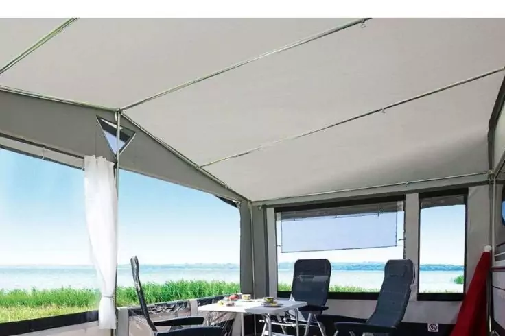 Innenraum eines Stoffvorbaus mit großen Fenstern zum Wasser, zwei Liegestühle, kleiner Tisch mit Geschirr und Vorhang links