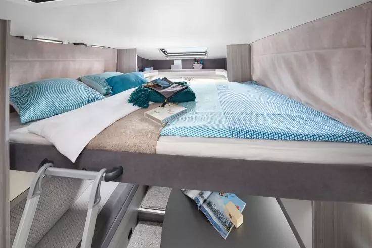 Hochbett mit blauer Bettwäsche, zwei Kissen und gefalteter Decke; Kleidung und Buch auf dem Bett; Leiter zum Bett; kleines Fenster und Leselicht; Regal unter dem Bett mit Magazin