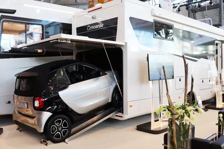 Weißes großes Wohnmobil mit geöffneter Heckgarage, in der ein kleines schwarz-weißes Stadtauto auf einer Rampe steht; im Vordergrund Pflanzen, im Hintergrund weitere Fahrzeuge und Hallenstruktur sichtbar
