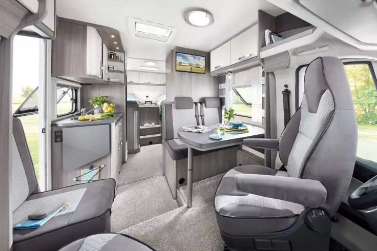 Innenraum eines Wohnmobils mit zwei vorderen Polstersitzen, Tisch mit Gedeck, Küchenzeile mit Spüle und Herd, Oberschränken, kleinem Wandfernseher und Fenstern mit Blick ins Grüne