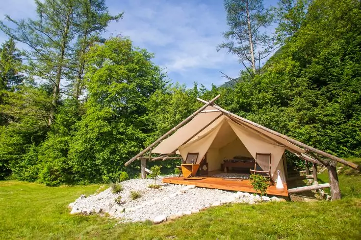 Beiges Zelt mit offenem Eingang auf Holzdeck, zwei Stühle und kleiner Tisch, weiße Kiesfläche davor, grüner Rasen und Bäume im Hintergrund, blauer Himmel mit Wolken
