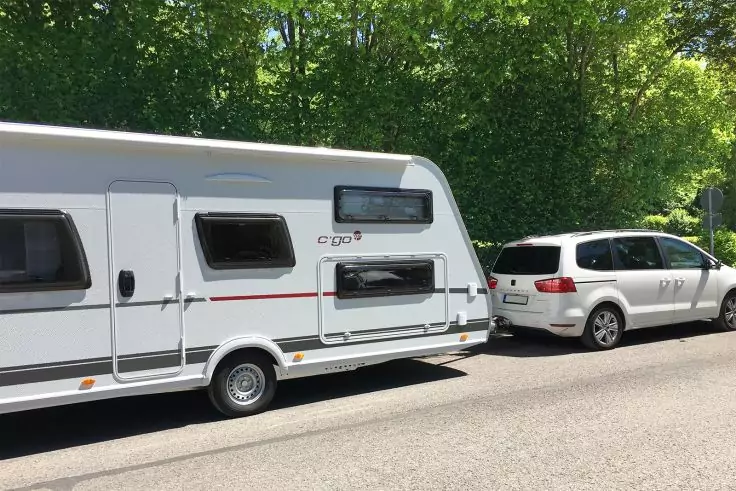 Weißer Kombi vor einem weißen Wohnwagen am Straßenrand, im Hintergrund grüne Bäume