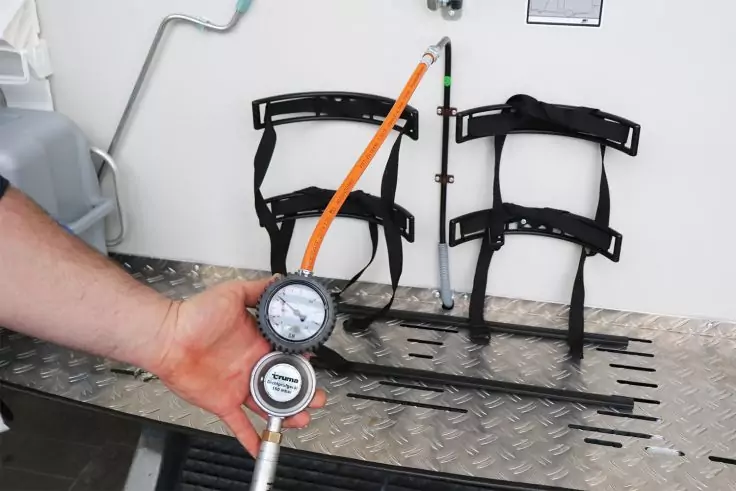 Hand hält zwei runde Messanzeigen an einem orangefarbenen Schlauch über einer Riffelblechfläche; an der weißen Wand zwei schwarze, gebogene Halterungen und mehrere flache Metallstreifen