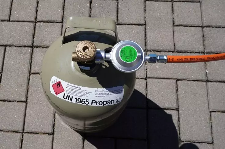 Beige Gasflasche mit angeschlossenem Druckregler und orangefarbenem Schlauch auf gepflastertem Boden, Etikett 'UN 1965 Propan' sichtbar