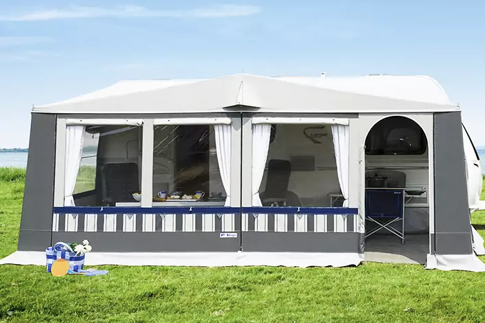 Großes weiß-graues Vorzelt an einem Caravan mit drei Frontfenstern und weißen Gardinen, Innenraum mit Stühlen und kleiner Campingküche sichtbar, grüner Rasen im Vordergrund, Wasser und blauer Himmel im Hintergrund