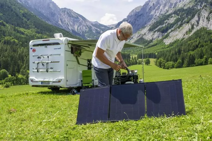 Mann mit weißem Haar stellt ein faltbares Solarmodul neben einem Wohnmobil auf, auf einer Wiese mit Bergen im Hintergrund