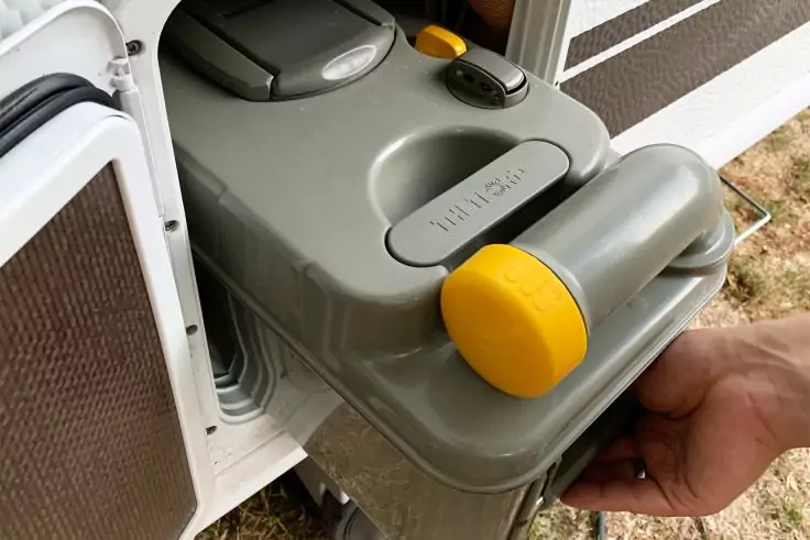 Hand zieht einen grauen Kunststoffbehälter mit zwei gelben runden Griffen aus einem weißen Fach; braunes Panel rechts, trockenes Gras unten